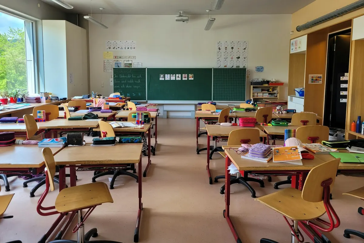 Aula de primaria luminosa, con pupitres de madera, pizarra verde y un gran ventanal a los árboles.