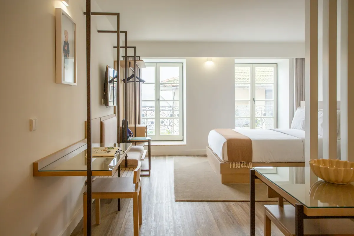 Habitación de hotel luminosa, con cama de matrimonio, escritorio de madera y dos balcones abiertos a la calle.