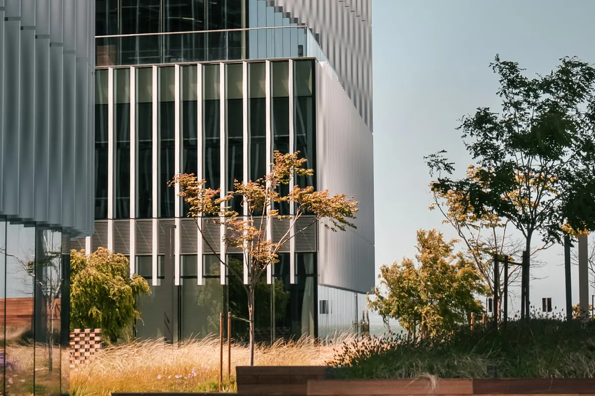 Fachada de un edificio de oficinas de vidrio y lamas metálicas, con un patio ajardinado.